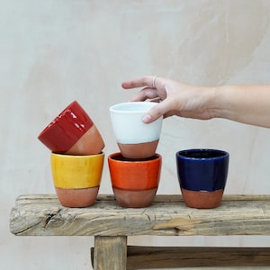 May include: A selection of brightly coloured ceramic cups with a two-tone design. The cups are in red, yellow, orange, white, and blue, with a terracotta base. They are displayed on a rustic wooden surface.