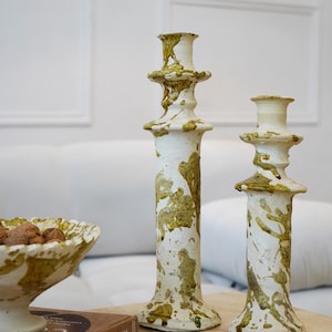 May include: Two white ceramic candlesticks with olive green splatter designs. A matching ceramic bowl with nuts sits next to the candlesticks. The items are on a wooden surface.
