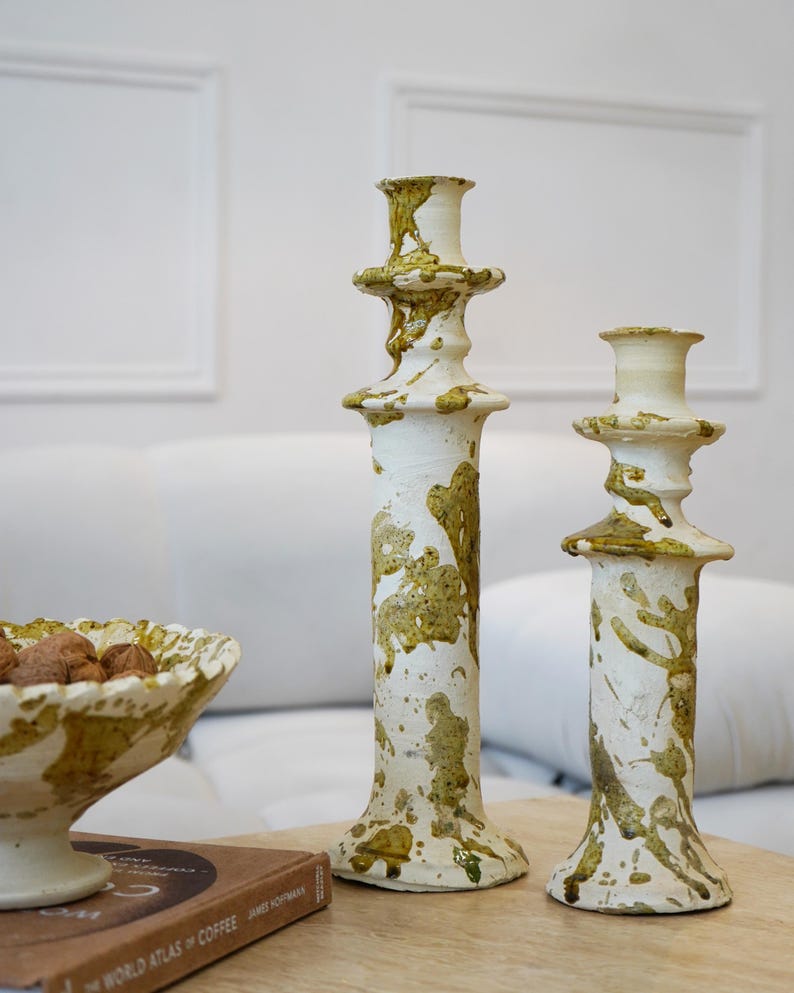 May include: Two white ceramic candlesticks with olive green splatter designs. A matching ceramic bowl with nuts sits next to the candlesticks. The items are on a wooden surface.