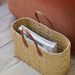 Op de afbeelding: Rechthoekige rieten mand met bruine leren handvatten, gevuld met tijdschriften. De mand is gemaakt van lichtbruine natuurlijke vezels. De handvatten zijn bevestigd met kleine leren patches. De mand staat op een lichtgekleurd tapijt.