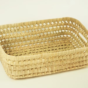 May include: Rectangular woven basket made of natural fibers. The basket has a rectangular shape with a woven pattern and small handles on the sides. The basket is a light tan color and appears to be handmade.