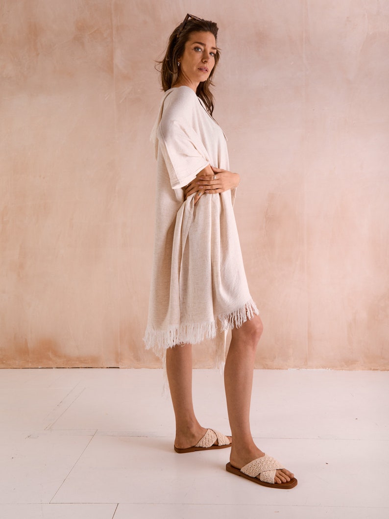 Puede incluir: Una bata de playa blanca de algod&oacute;n con detalles de flecos. La bata se lleva sobre un traje de ba&ntilde;o y tiene un corte holgado.