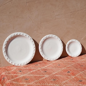 May include: Three white ceramic plates of varying sizes, each with a beaded rim. The plates are arranged on a textured, peach-colored rug, with a solid peach-colored wall in the background. The plates are in direct sunlight.