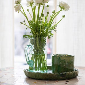 Op de afbeelding: Een heldere glazen vaas met een handvat bevat een boeket witte bloemen met groene stelen. Een groene keramische kandelaar en een bijpassend dienblad staan naast de vaas. De items staan op een mozaïek tegel tafel.