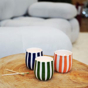 May include: Three ceramic candle holders with vertical stripes in blue, green, and orange, each with a white interior. The candle holders are on a wooden surface, with two matches nearby. The background is blurred.