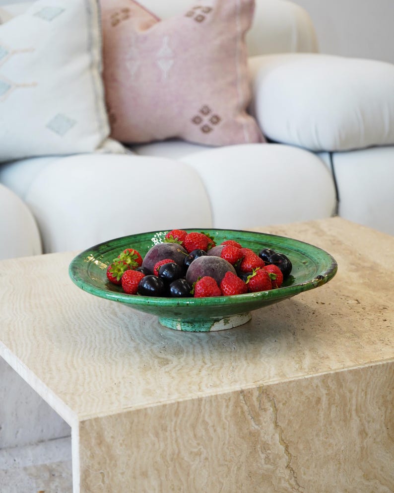 May include: A green ceramic bowl filled with fresh fruit, including strawberries, blueberries, and peaches, sits on a light-colored stone table. The bowl has a wide rim and a pedestal base. In the background, a white sofa and decorative pillows are visible.