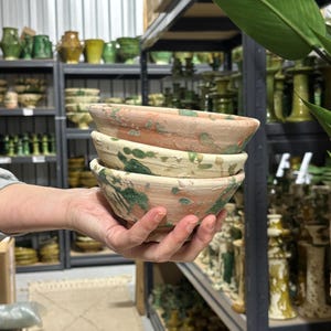 Może przedstawiać: Stos trzech ceramicznych misek o rustykalnym, postarzanym wyglądzie. Miski mają kremową podstawę z nakrapianymi zielonymi i pomarańczowymi akcentami. Miski są trzymane na tle półek wypełnionych ceramiką.