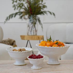 May include: Three white ceramic pedestal bowls with scalloped edges of varying sizes. The largest bowl is filled with bright orange tangerines, the medium bowl contains brown walnuts, and the smallest bowl holds a cluster of red currants.