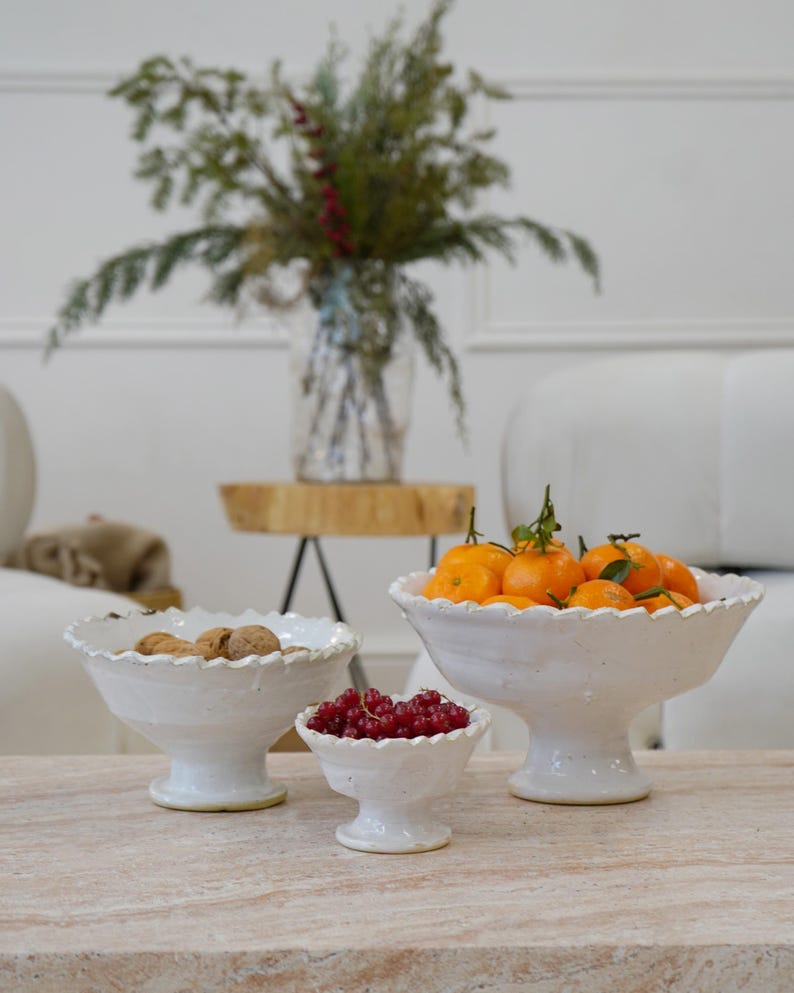 May include: Three white ceramic pedestal bowls with scalloped edges of varying sizes. The largest bowl is filled with bright orange tangerines, the medium bowl contains brown walnuts, and the smallest bowl holds a cluster of red currants.
