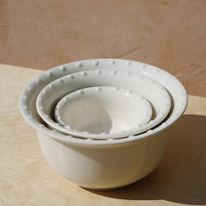 May include: A set of three white ceramic mixing bowls nested together. The bowls have a simple, elegant design with a scalloped rim and small, raised dots around the edge. The bowls are arranged on a light-colored wooden surface.