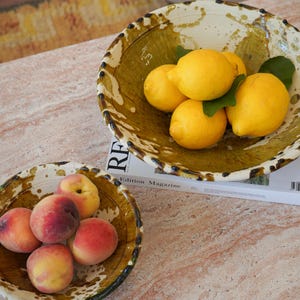 Può includere: Due ciotole rotonde in ceramica con un motivo a schizzi color crema e verde oliva. La ciotola più grande contiene quattro limoni giallo brillante con foglie verdi, mentre la ciotola più piccola contiene cinque pesche fresche dai toni rosa.
