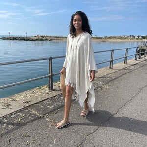 Puede incluir: Una mujer lleva un poncho blanco con capucha y detalles de flecos mientras camina por un paseo pavimentado junto al agua. Lleva un bolso de paja tejido.