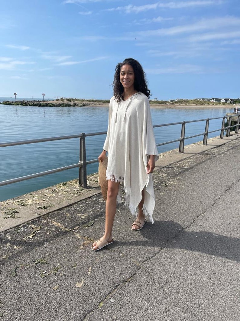 Puede incluir: Una mujer lleva un poncho blanco con capucha y detalles de flecos mientras camina por un paseo pavimentado junto al agua. Lleva un bolso de paja tejido.