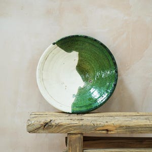 Puede incluir: Un plato de cer&aacute;mica hecho a mano con un dise&ntilde;o &uacute;nico. El plato es mitad blanco y mitad verde, con un acabado brillante. El plato est&aacute; sobre un banco de madera r&uacute;stica. Es un objeto decorativo.