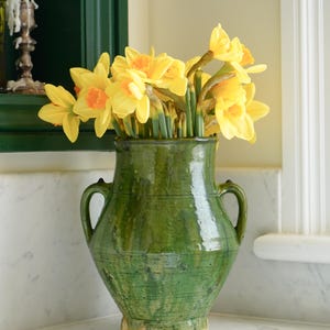 May include: A green ceramic vase with two handles holds a bouquet of yellow daffodils with orange centers. The vase has a textured surface and sits on a white marble surface. The background includes a dark green window frame and a white trim.