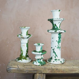 May include: Three ceramic candle holders of varying heights. The candle holders are white with green paint splatters. They are arranged on a weathered wooden surface against a neutral background.