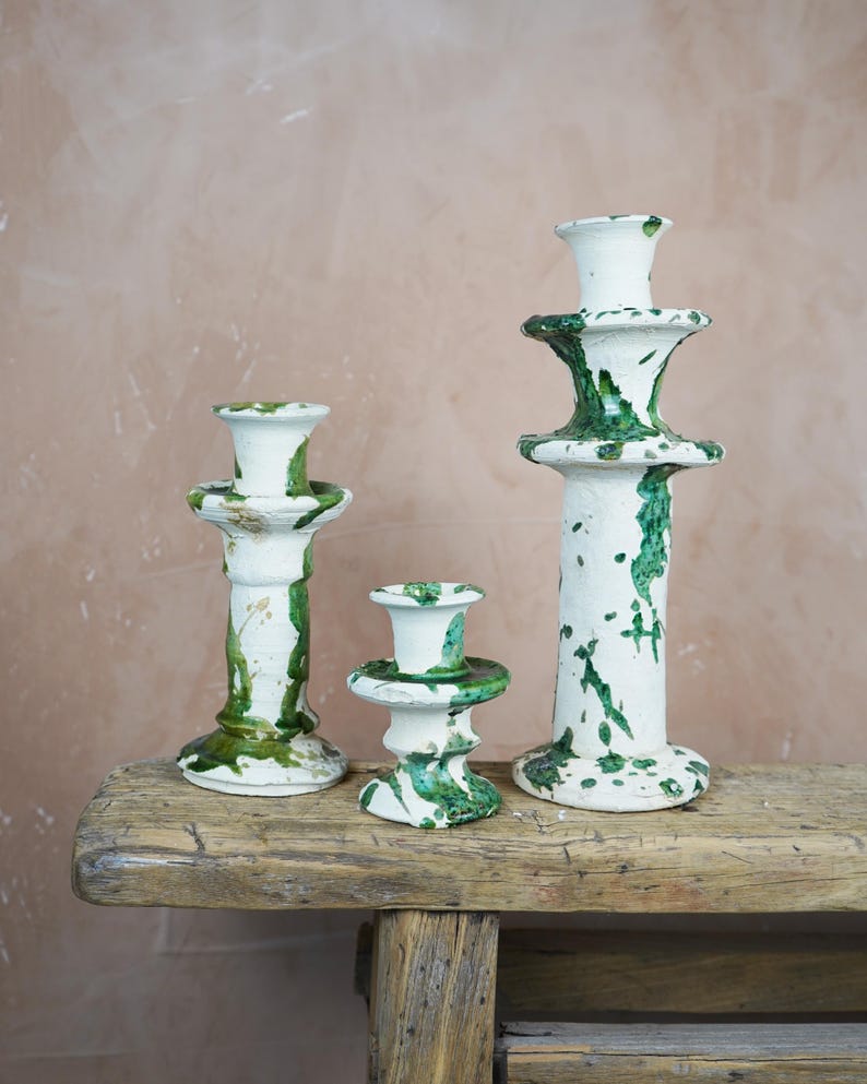 May include: Three ceramic candle holders of varying heights. The candle holders are white with green paint splatters. They are arranged on a weathered wooden surface against a neutral background.