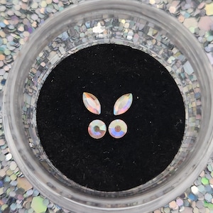May include: A small clear container filled with iridescent glitter. Inside the container, there are five iridescent rhinestones, two oval-shaped and three round.