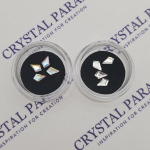 Op de afbeelding: Twee kleine doorzichtige plastic containers met zwarte deksels, elk met een set iriserende, gefacetteerde, geometrisch gevormde strass steentjes. De containers zijn gelabeld met "Crystal Paradise Inspiration for Creation".