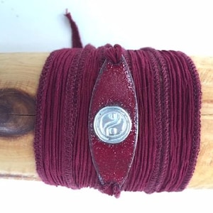 May include: A burgundy wrap bracelet with a silver yin yang charm. The bracelet is made of multiple strands of cord and is tied with a knot.