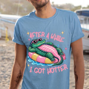 May include: Light blue t-shirt featuring a cartoon alligator wearing sunglasses and floating on a pink and yellow inflatable ring. The text "AFTER A WHILE I GOT HOTTER" is printed above and below the alligator.