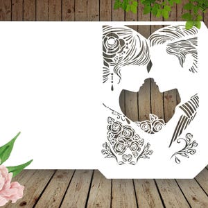 K&ouml;nnte beinhalten: Eine wei&szlig;e, lasergeschnittene Hochzeitseinladungskarte mit einer Silhouette eines sich k&uuml;ssenden Paares. Das Design beinhaltet florale und dekorative Elemente. Die Karte befindet sich vor einem Holzgrund mit Aquarellblumen.