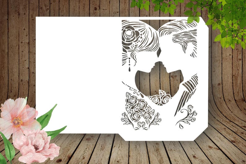 K&ouml;nnte beinhalten: Eine wei&szlig;e, lasergeschnittene Hochzeitseinladungskarte mit einer Silhouette eines sich k&uuml;ssenden Paares. Das Design beinhaltet florale und dekorative Elemente. Die Karte befindet sich vor einem Holzgrund mit Aquarellblumen.