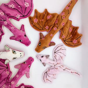 May include: A collection of four plush dragon toys in pink, brown, white, and light pink. Each dragon features embroidered cherry blossom patterns on its wings and body, with detailed stitched eyes and small horns on their heads.