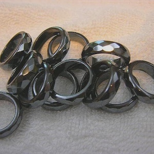May include: A collection of dark gray hematite rings. Some rings have a faceted design, while others are smooth. The rings are displayed on a white, textured surface. The rings vary in size and shape.