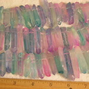 Könnte beinhalten: Eine Reihe von regenbogenfarbenen Quarzkristallspitzen in Rosa-, Blau- und Grüntönen. Die Kristalle sind etwa 2,5 cm lang und in einer Reihe auf einer weißen Oberfläche angeordnet. Ein Lineal ist im unteren Teil des Bildes sichtbar.