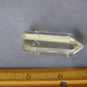 Puede incluir: Una punta de cristal de cuarzo transparente, de aproximadamente 5 cm de largo, con forma hexagonal y punta puntiaguda. El cristal está sobre una superficie azul.