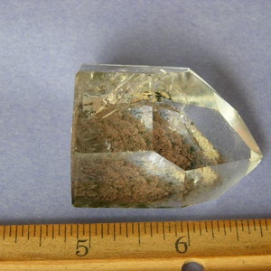 Puede incluir: Un cristal de cuarzo transparente con inclusiones de minerales marrones y blancos. El cristal está facetado y tiene una punta puntiaguda.