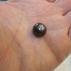 May include: A small, spherical object with a dark, metallic, and reflective surface. The item has a smooth, polished finish that catches the light, creating a bright highlight on its rounded top. The object is solid and uniform in its dark grey colour.