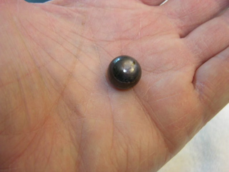 May include: A small, spherical object with a dark, metallic, and reflective surface. The item has a smooth, polished finish that catches the light, creating a bright highlight on its rounded top. The object is solid and uniform in its dark grey colour.