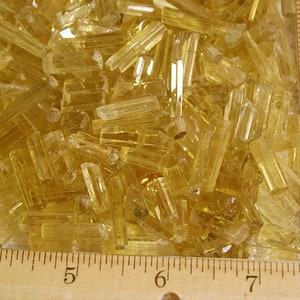 Peut inclure: Gros plan sur un tas de petits cristaux translucides jaune doré. Les cristaux sont disposés de manière aléatoire et mesurent environ 2,5 cm de long. Une règle est visible en arrière-plan, avec des marques de 10 à 18 cm.