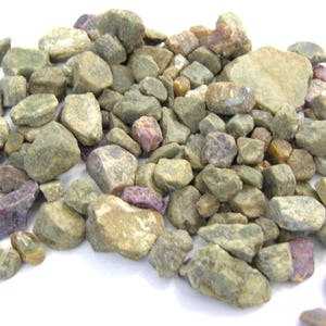 May include: A pile of rough-cut gemstones in shades of green, tan, and purple. The stones vary in size and shape, with a slightly matte finish. The gemstones are scattered on a white surface.
