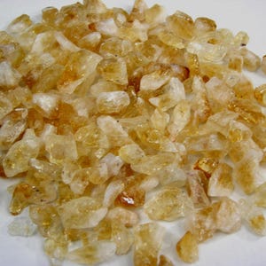 Puede incluir: Un montón de cristales de citrino en bruto. Los cristales son una mezcla de translúcidos y opacos, con colores que van del amarillo pálido al ámbar oscuro. Las piedras en bruto tienen formas irregulares y tamaños variables.