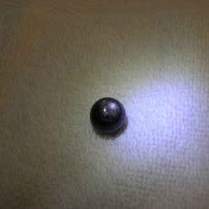 May include: A single, spherical, dark-coloured gemstone with a smooth, polished surface. The stone exhibits a distinct, star-like light reflection pattern on its top, characteristic of asterism, with a deep charcoal or black base colour.
