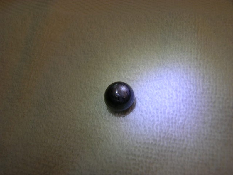 May include: A single, spherical, dark-coloured gemstone with a smooth, polished surface. The stone exhibits a distinct, star-like light reflection pattern on its top, characteristic of asterism, with a deep charcoal or black base colour.