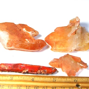 May include: Three orange and red iron-stained quartz crystal clusters and a single elongated red crystal point. A wooden ruler is positioned at the bottom, showing the longest crystal measures approximately 4.5 inches in length.