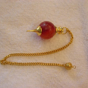 Peut inclure: Un pendule à chaîne dorée avec une sphère rouge et blanche mouchetée. Le pendule a une pointe pointue et une petite boule dorée au bout de la chaîne.