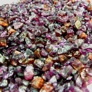 May include: A close-up view of a pile of rough-cut gemstones. The stones are primarily shades of purple, with hints of orange and brown. The gemstones have a crystalline appearance and varying sizes, creating a textured surface.