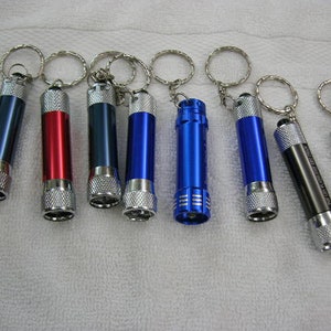 May include: A set of nine keychain flashlights in various colors, including blue, red, and black. Each flashlight has a silver metal ring for attaching to a keychain.