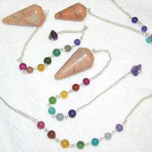 Op de afbeelding: Set van zes roze en witte kristallen pendels met zilveren kettingen en gekleurde kralen. Elke pendel heeft een ketting met kralen in een andere kleur, waaronder rood, oranje, geel, groen, blauw en paars.