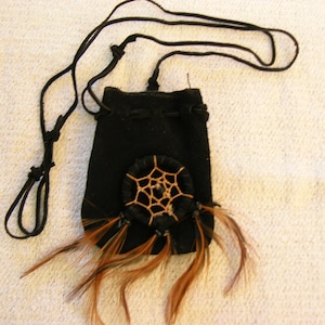 May include: A small black leather pouch with a brown dreamcatcher design and feathers hanging from the bottom. The pouch has a black cord for wearing it as a necklace.