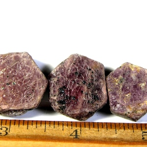 May include: Three raw, hexagonal-shaped ruby crystals with deep magenta and dark purple hues, positioned above a wooden ruler showing a scale in inches, with the stones spanning from the three-inch mark to just past the five-inch mark.