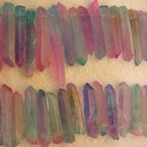 May include: Rows of iridescent quartz crystals in shades of pink, blue, and purple. The crystals are elongated and pointed, with a translucent quality. The arrangement suggests they could be used for jewelry or decorative purposes.