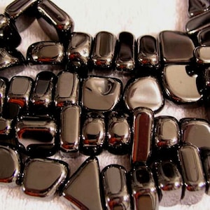 May include: A close-up of a necklace composed of polished, dark grey, rectangular and rounded stones. The stones are arranged closely together, reflecting light and creating a shiny appearance. The necklace is displayed on a white surface.