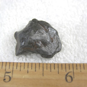 Puede incluir: Una piedra gris oscura de forma irregular con una textura rugosa. La piedra está colocada sobre una superficie blanca junto a una regla de madera. La regla muestra medidas en centímetros.