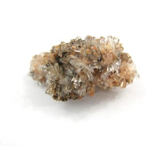 May include: A cluster of small, translucent crystal formations with a light brown and tan color palette. The crystals are clustered together, creating a textured appearance. The mineral specimen is set against a white background.